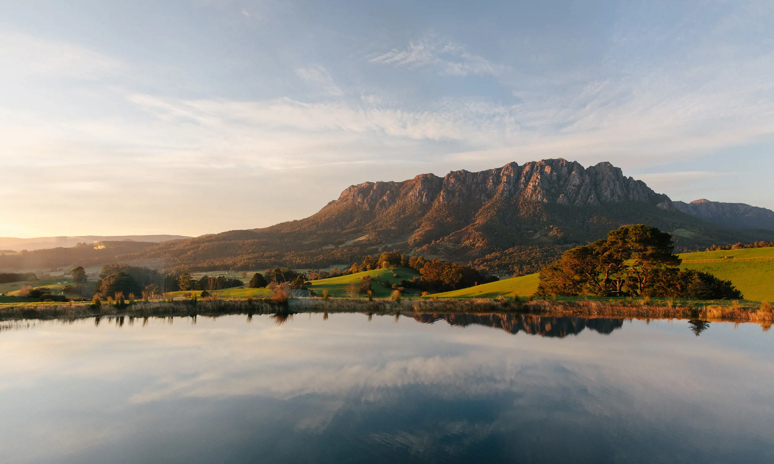 A picturesque landscape, looking over a glassy lake with the cloud-streaked sky reflected. Behind, a large mountain rises up over rolling green hills.