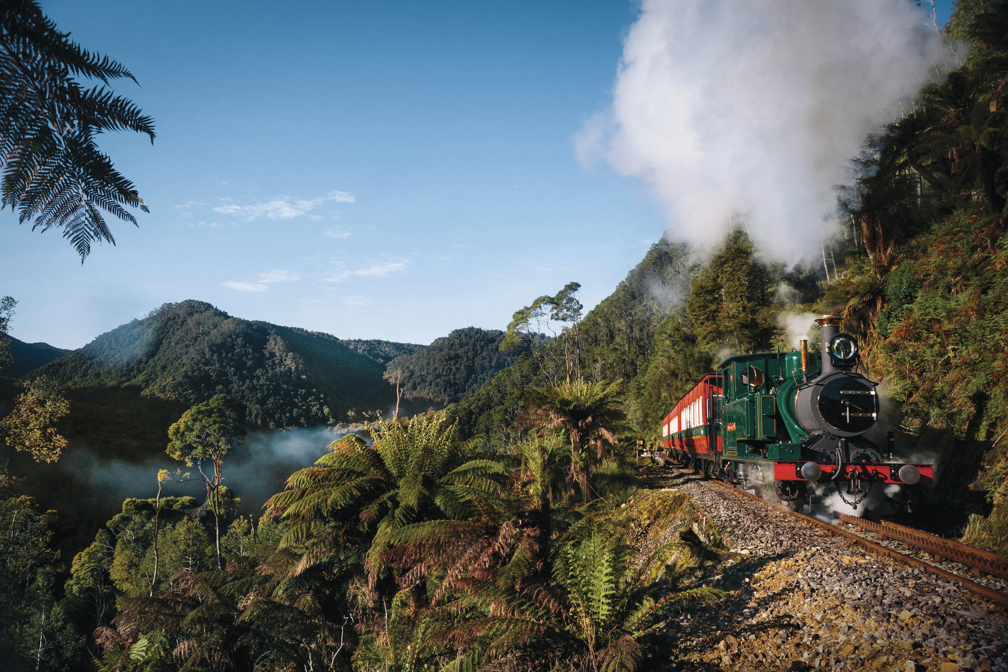 A green heritage steam engine chugs along tracks on the side of a steep hill, with thick bush, mountains and a river nearby.