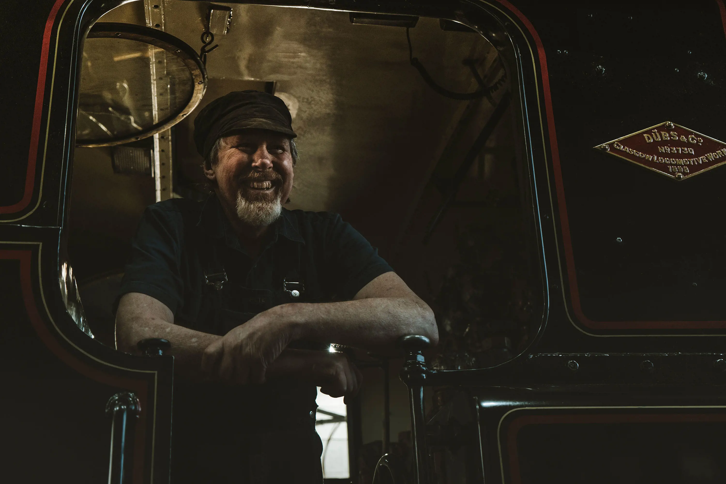 A bearded man in a black shirt and flatcap smiles down from the driver's cab of a shiny black heritage train.