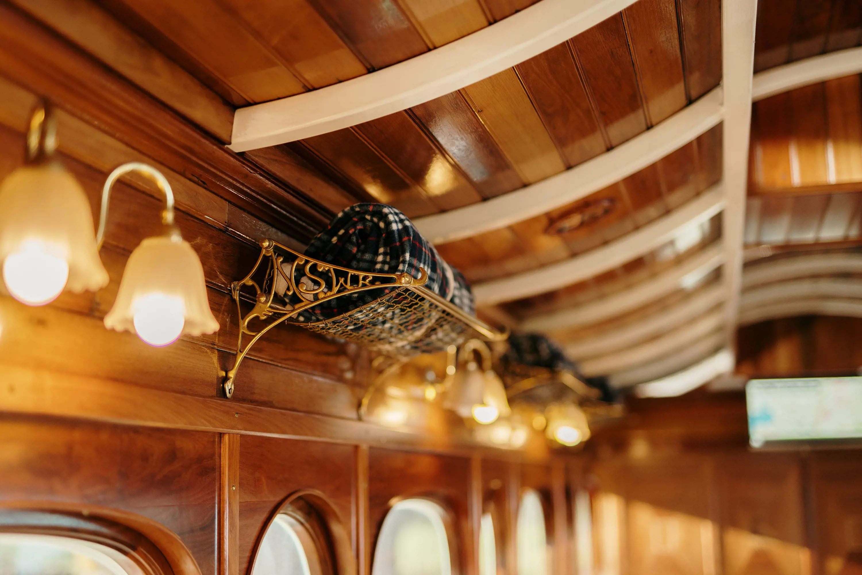 The rich wood interior of a heritage railway carriage, with decorative sconce lights and polished gold luggage racks with the letters 'NSWR' holding a picnic blanket.