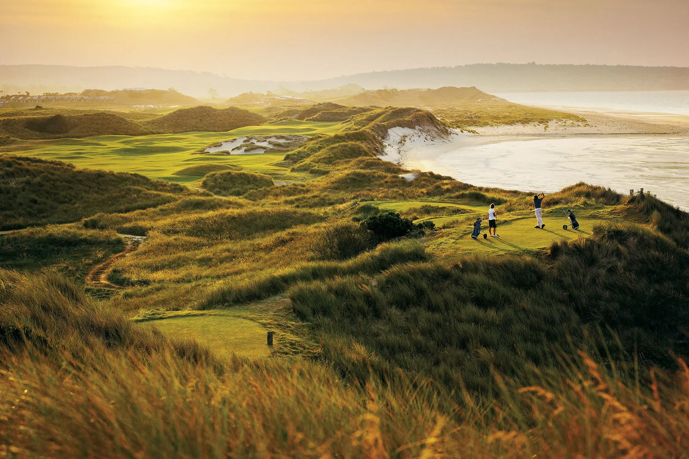 Looking across a landscape lit by sunset, dotted throughout with golf greens and rolling beachside dunes. The coast curves around to reveal a small bay.