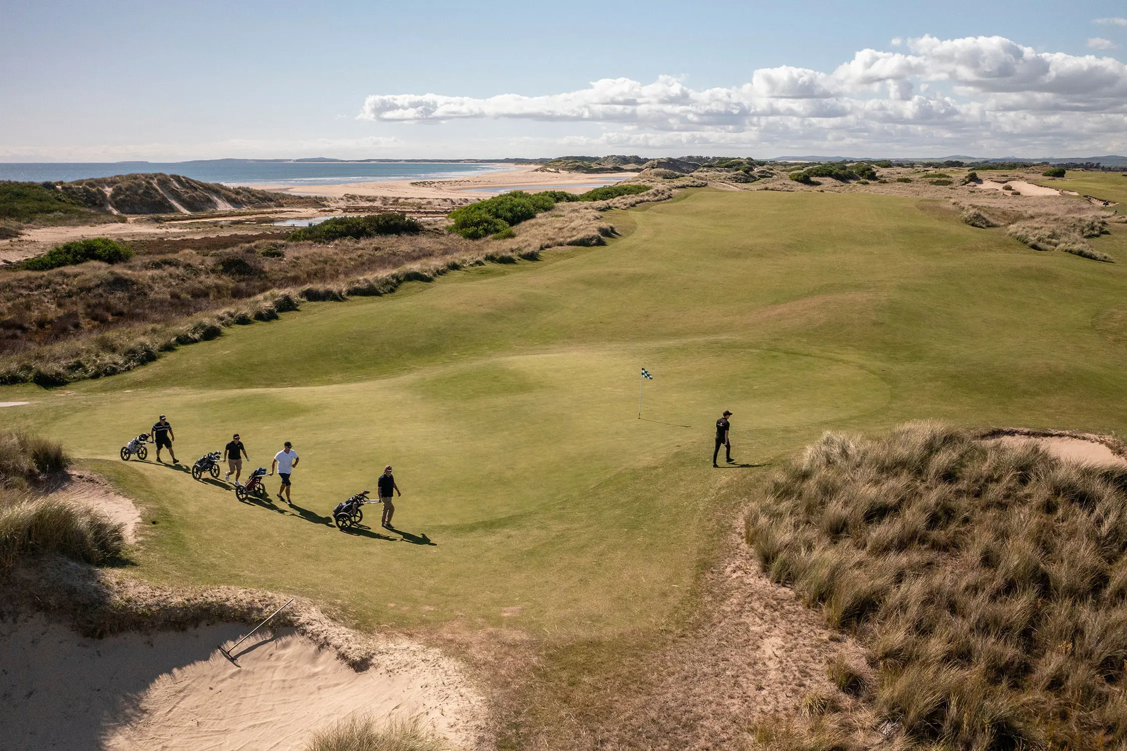 An aerial view of a rolling golf green surrounded by beach dunes and landscape, where five golfers walk with their carts.