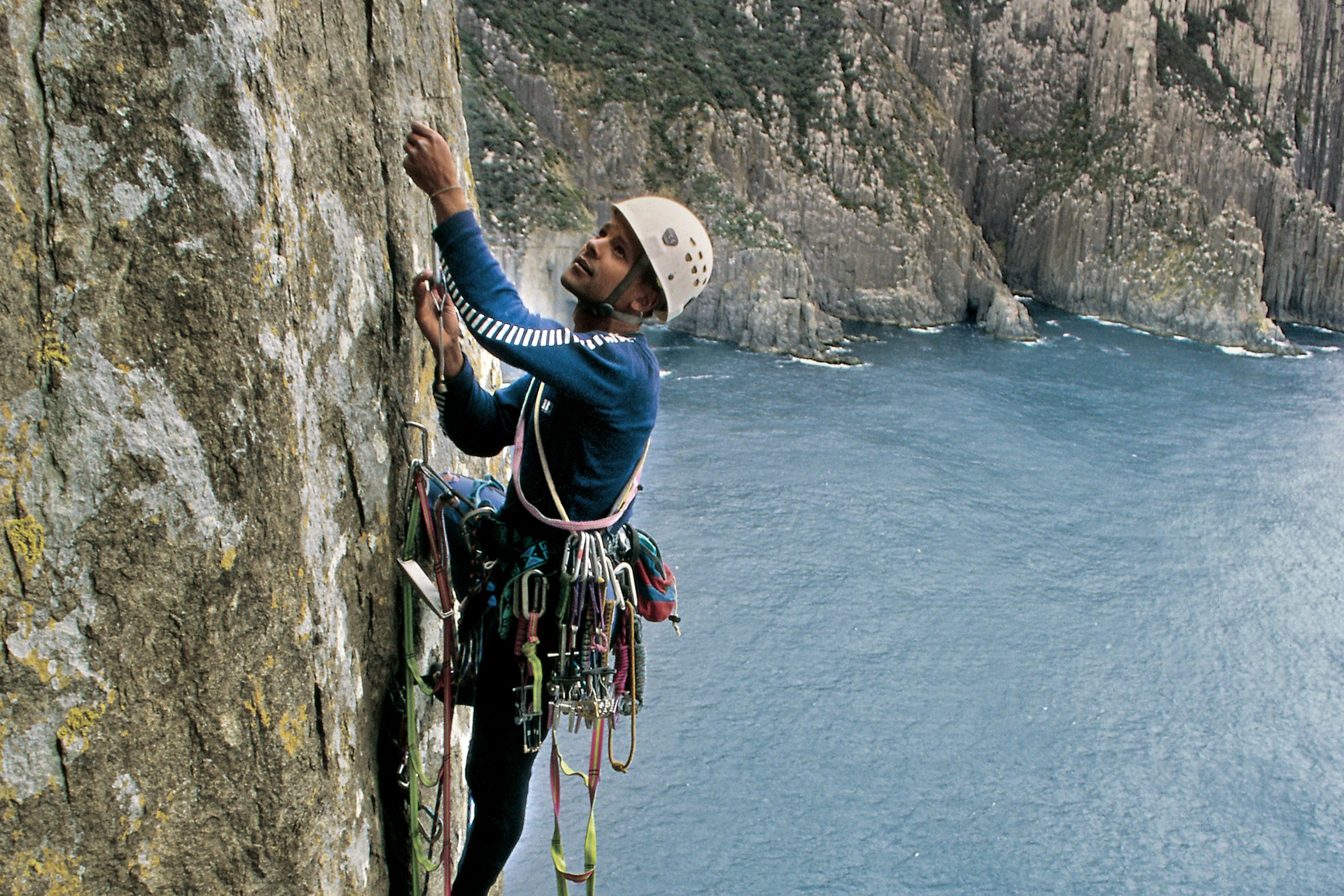 Jaw Dropping image of a person rock climbing up Totem Pole, Cape Hauy. A stunning landscape of mountains and ocean surround them.