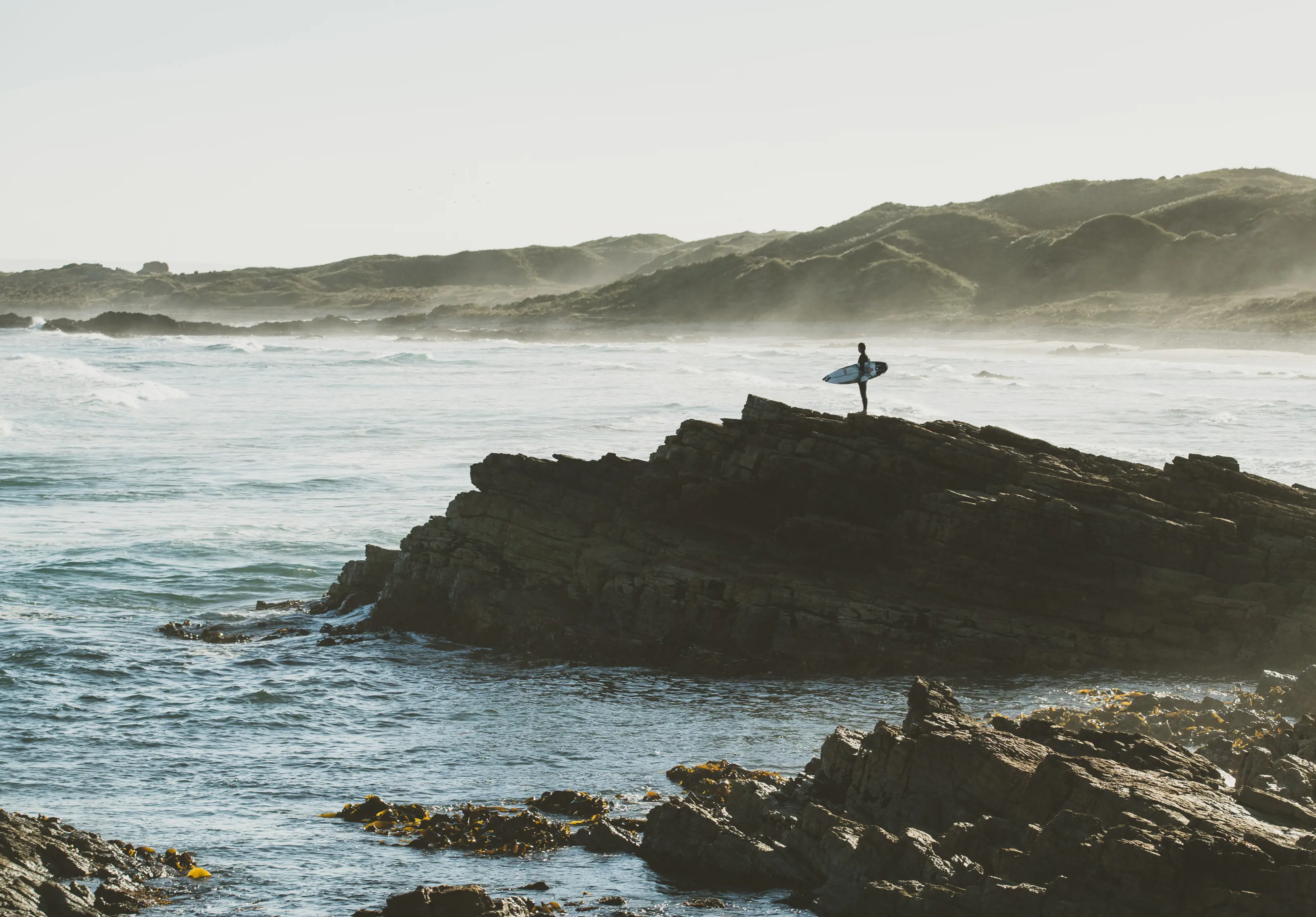 Dramatic image of a surfer standing on the edge of the rocks looking out to the ocean on sunrise.