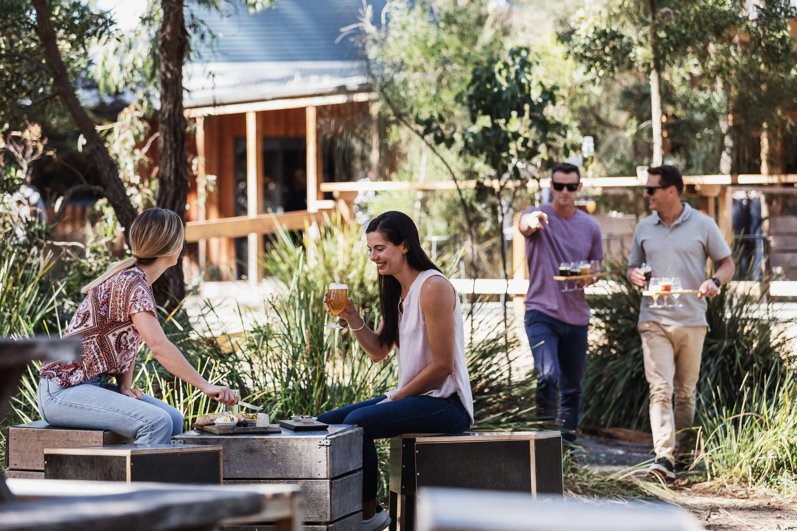 Two women enjoying a drink and cutting cheese while two men walk over to join them with an assortment of beers at Bruny Island Cheese Co.