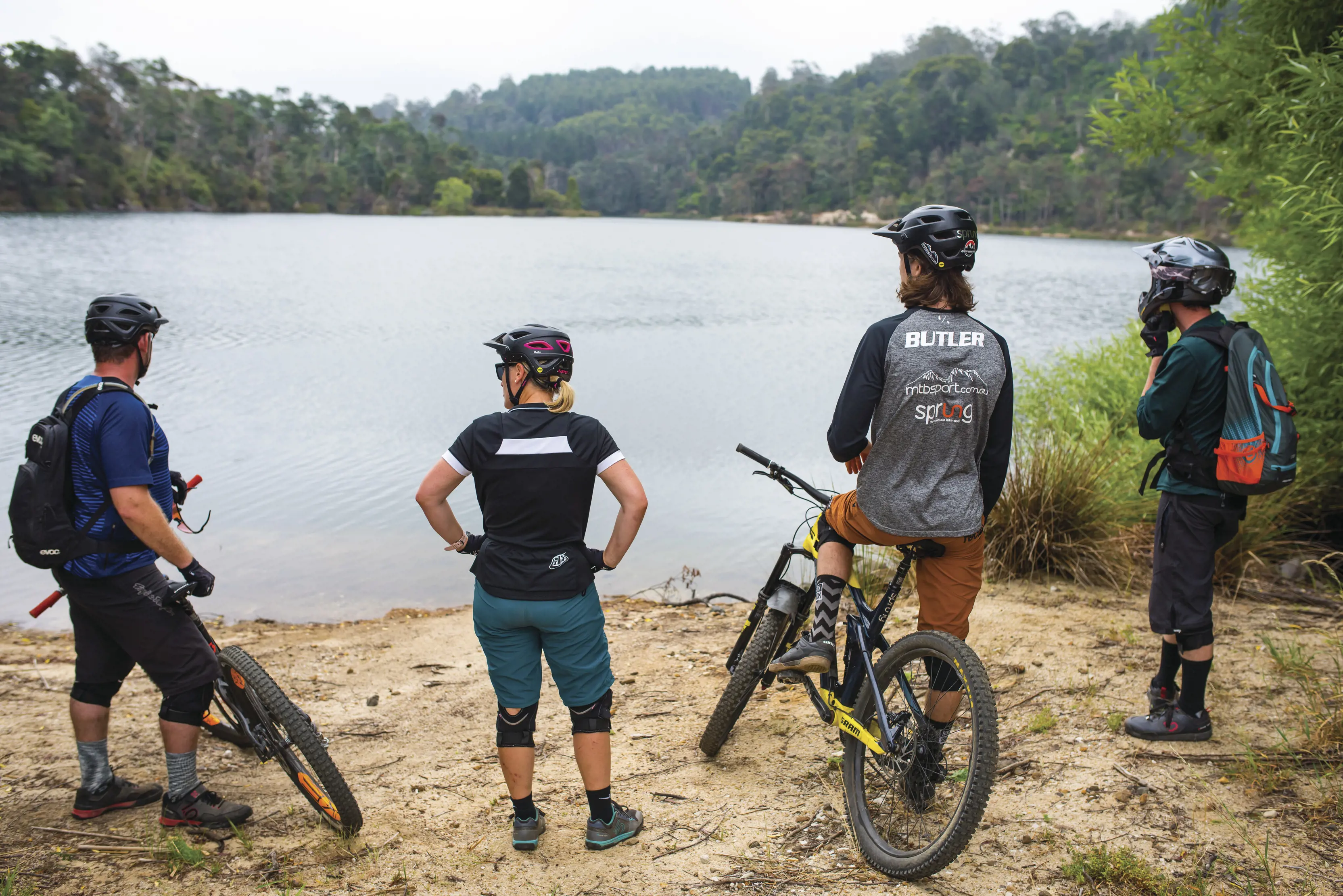 Four bike riders taking a break along a river bank, looking out into the wilderness, with Blue Derby Mountain Bike Trails.