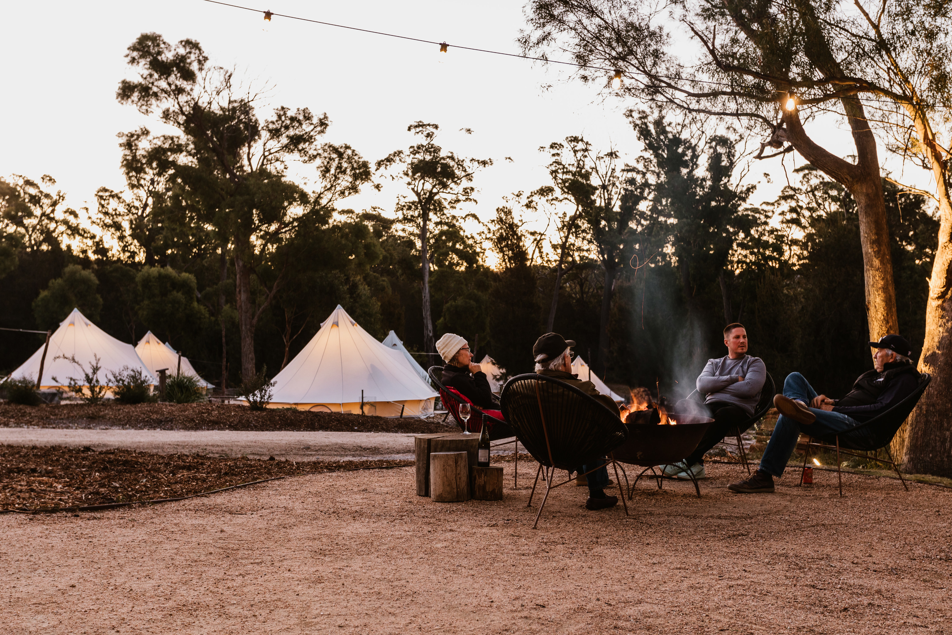 Four people sitting around a camp fire at Bay of Fires Bush Retreat. Pitched, white tents in the background