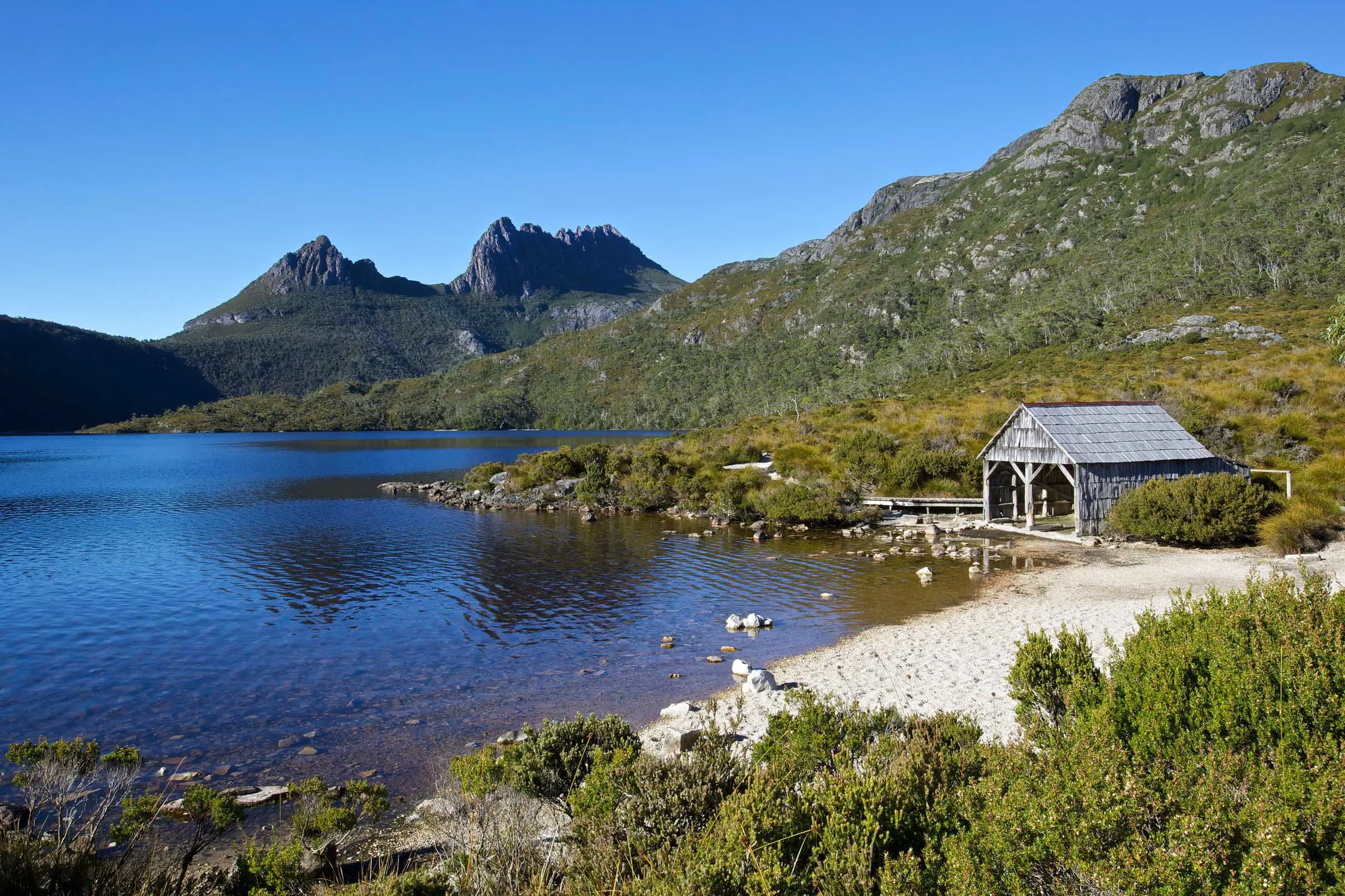 A small wooden boat shed sits on the sandy foreshore of a dark lake surrounded by bush. In the background, the sharp peaks of Cradle Mountain rise up.