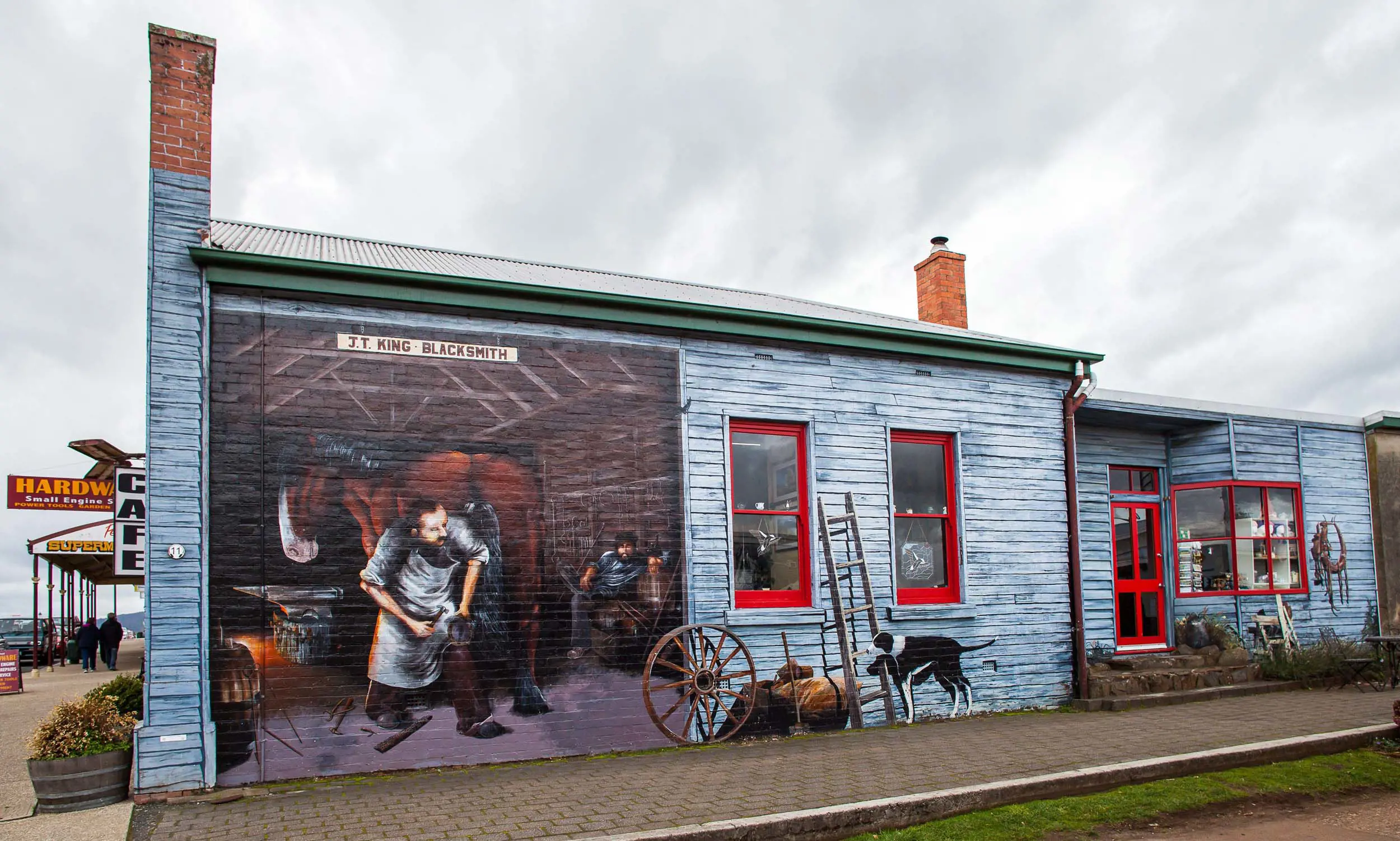 The side of a brick building is painted intricately to appear like a blue weatherboard building, with scenes of a blacksmith's inside.