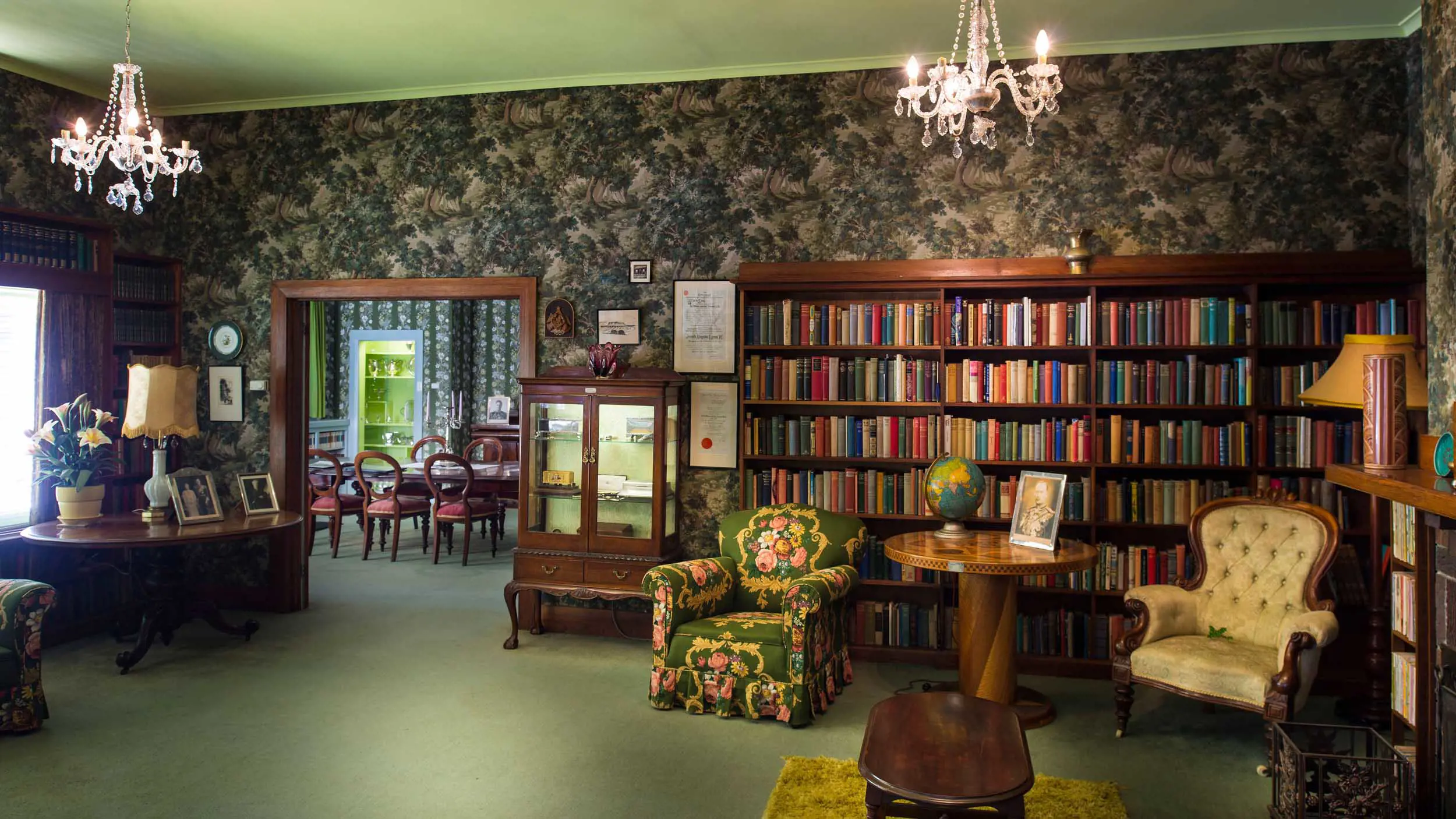 A vintage library room with bookshelves, floral wallpaper, chandeliers, antique furniture, and a globe on a table. A view through the doorway shows another room with a dining table and chairs.