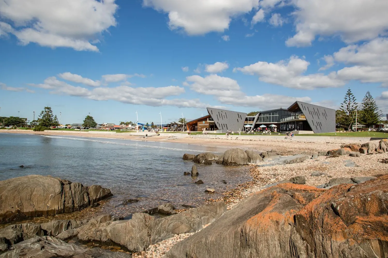 From some rocks to the side of a shallow beach, looking out to the shore where people sit on a grassed area around angular architectural buildings.