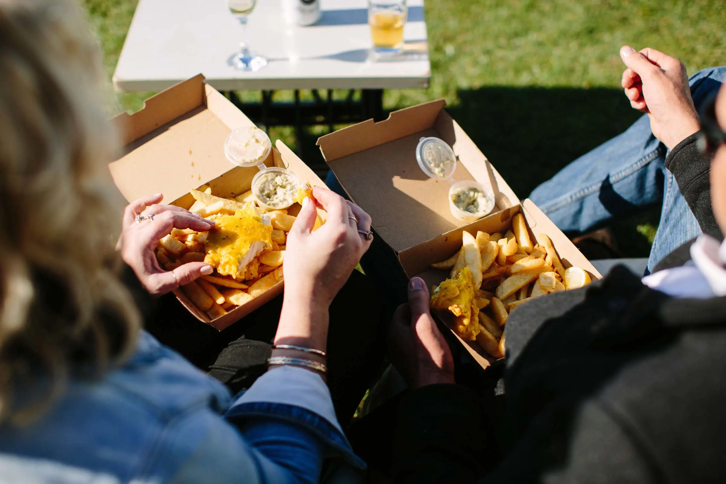 Looking over two people's shoulders as they sit on a bench, with two cardboard takeaway boxes full of fish and chips sitting on their laps.