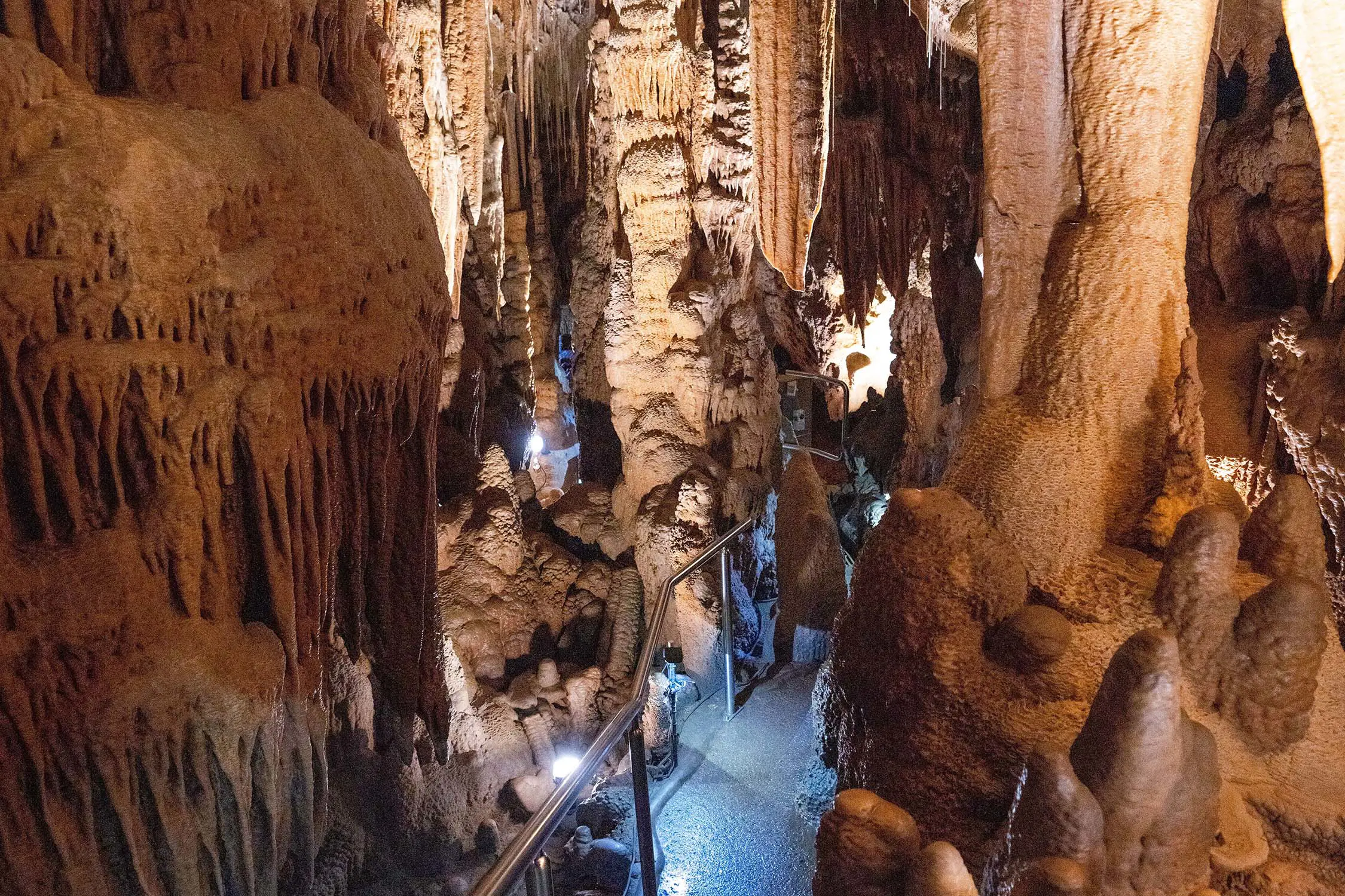 A path with a metal handrail through an underground cave, filled with tall stalagmites and stalactites, and other orange-brown rock formations.