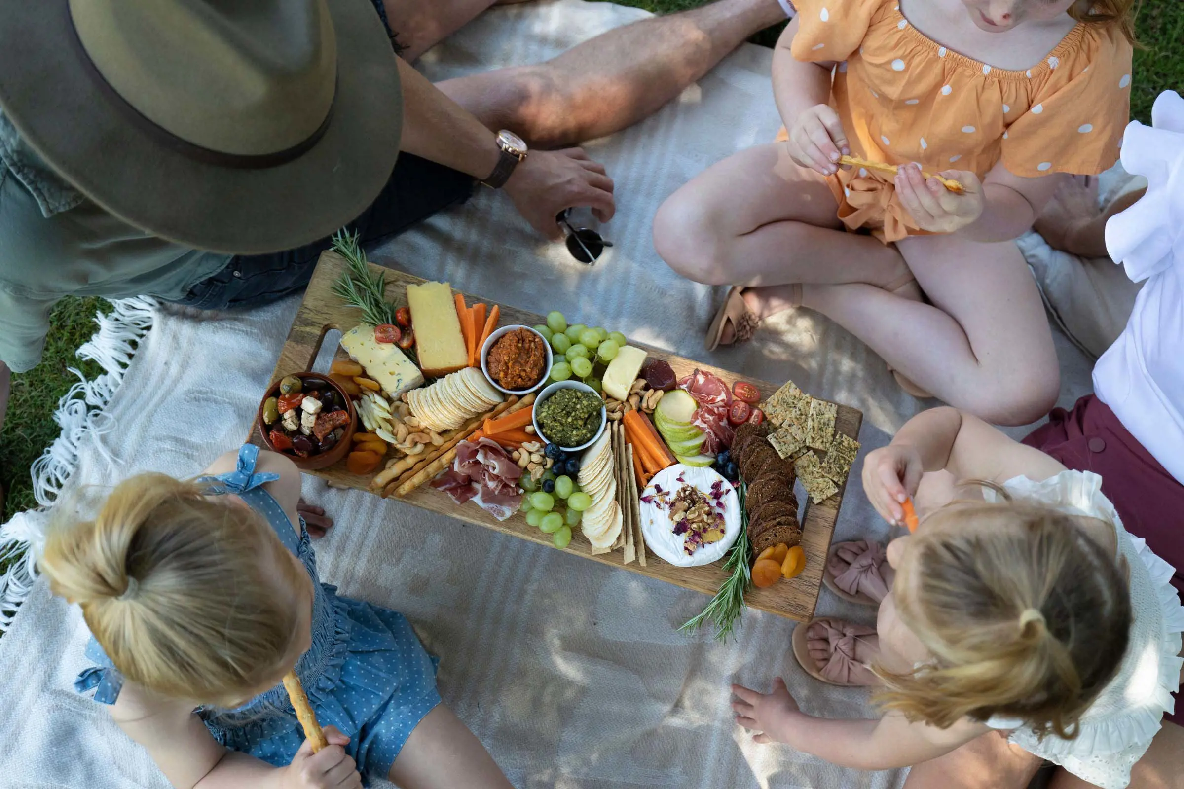 Looking top-down onto a delicious, stacked grazing platter on a picnic blanket. Three kids and two adults sit around it, reaching for something to eat.