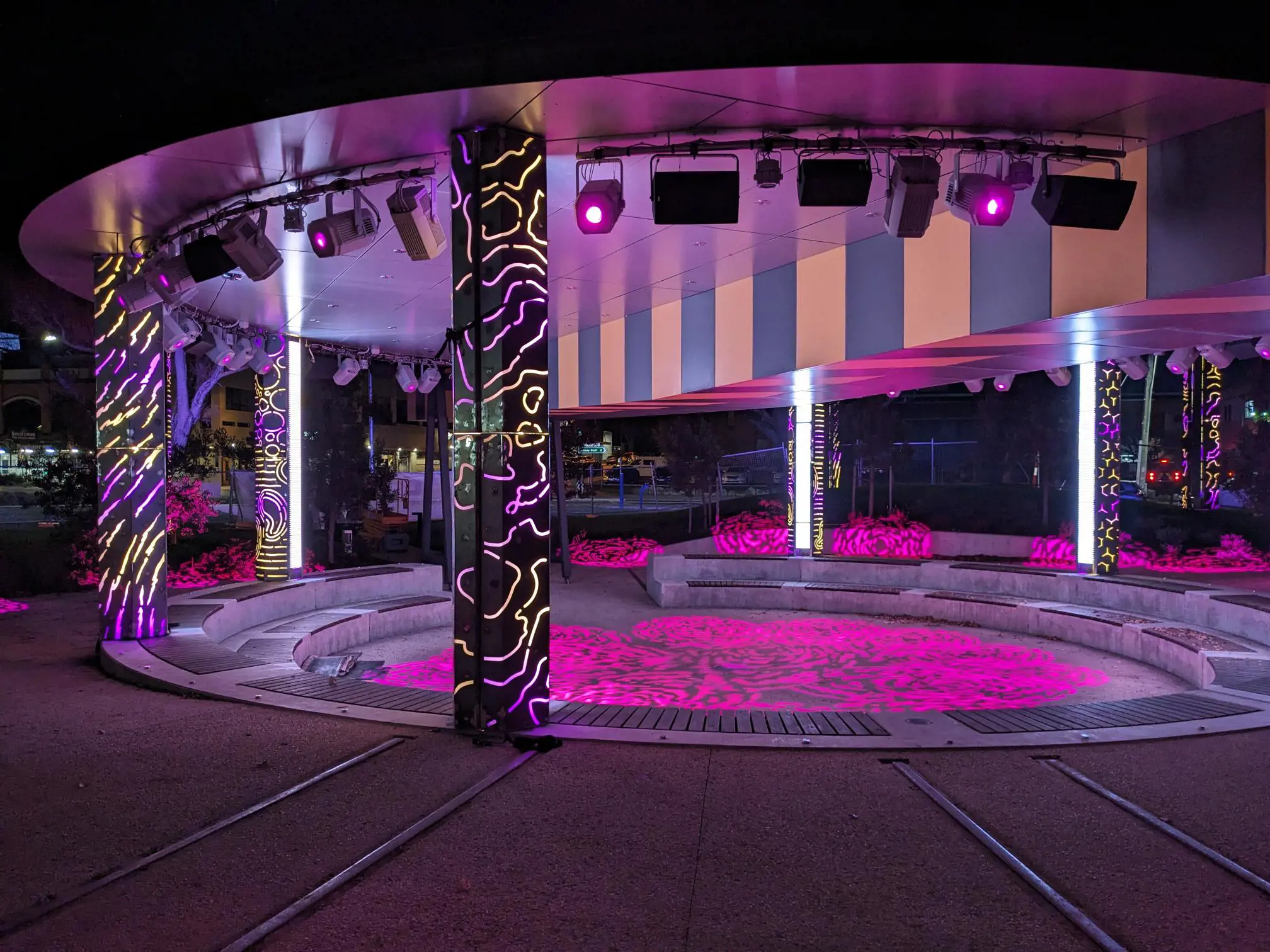 An outdoor art installation at night. Colourful purple and pink lights illuminate the circular overhead structure, with rows of seats surrounding the sunken centre, amphitheatre-style.