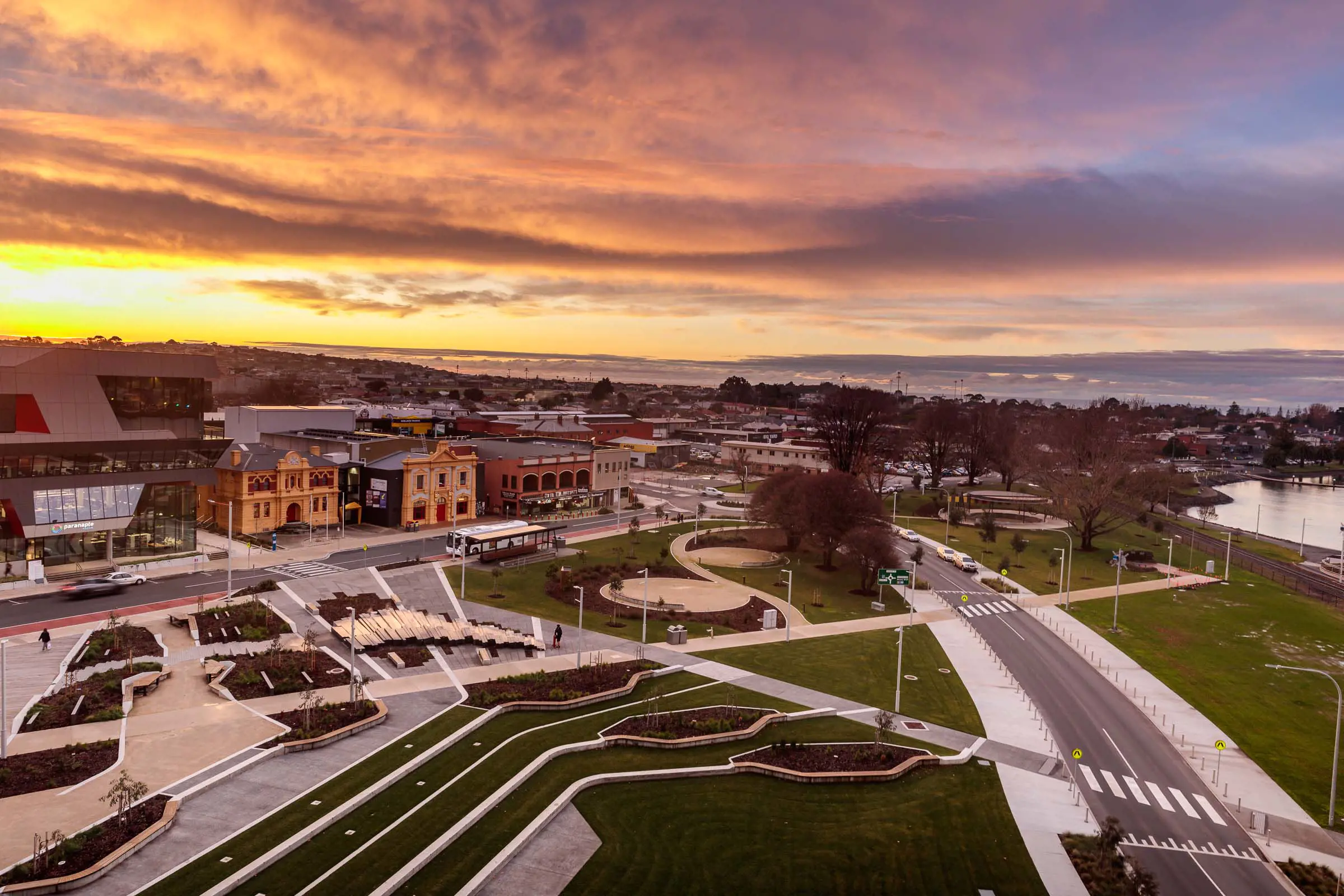 An aerial view of a waterfront development, featuring multiple footpaths, tiered seating areas, trees, bike paths and roads, streetlights and a mix of grassed, sandy and paved areas.