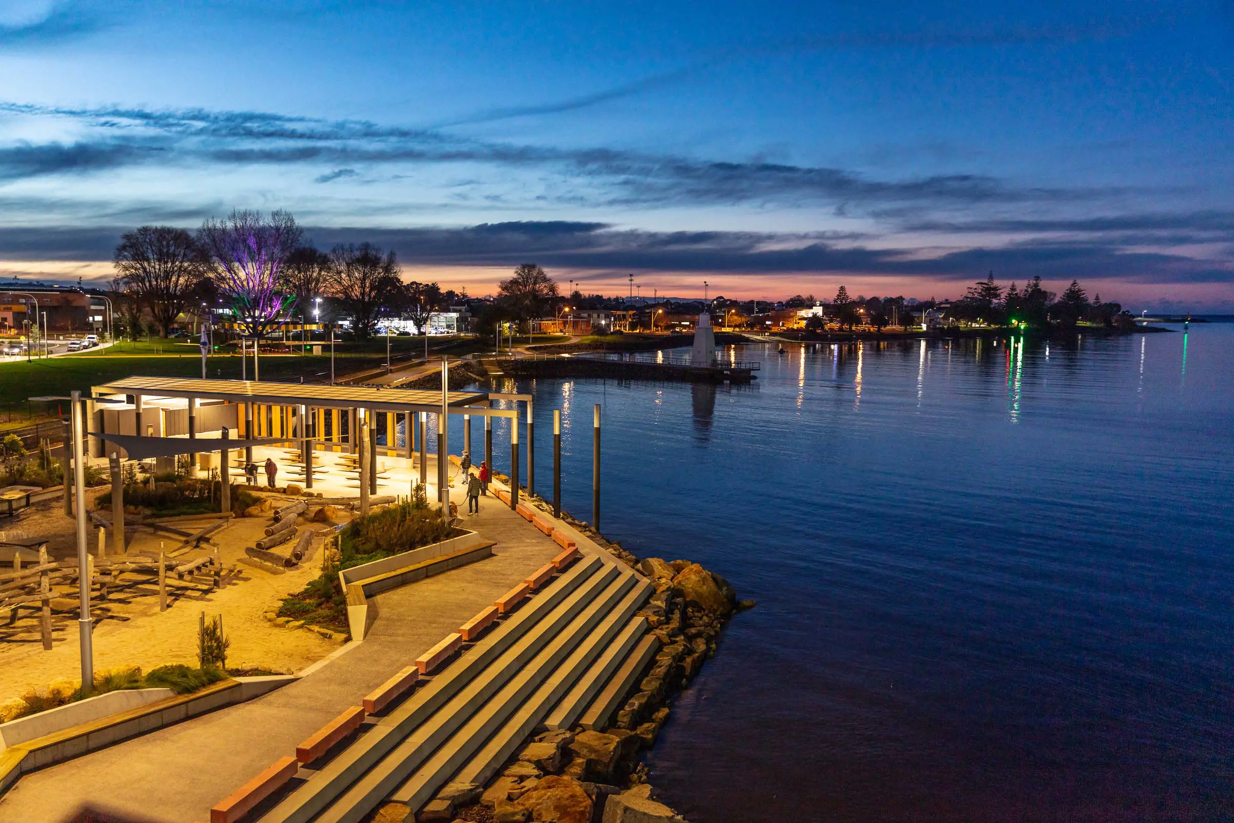 A waterfront area at twilight, with seating areas and steps down to the water. The lights of Devonport reflect in the water.