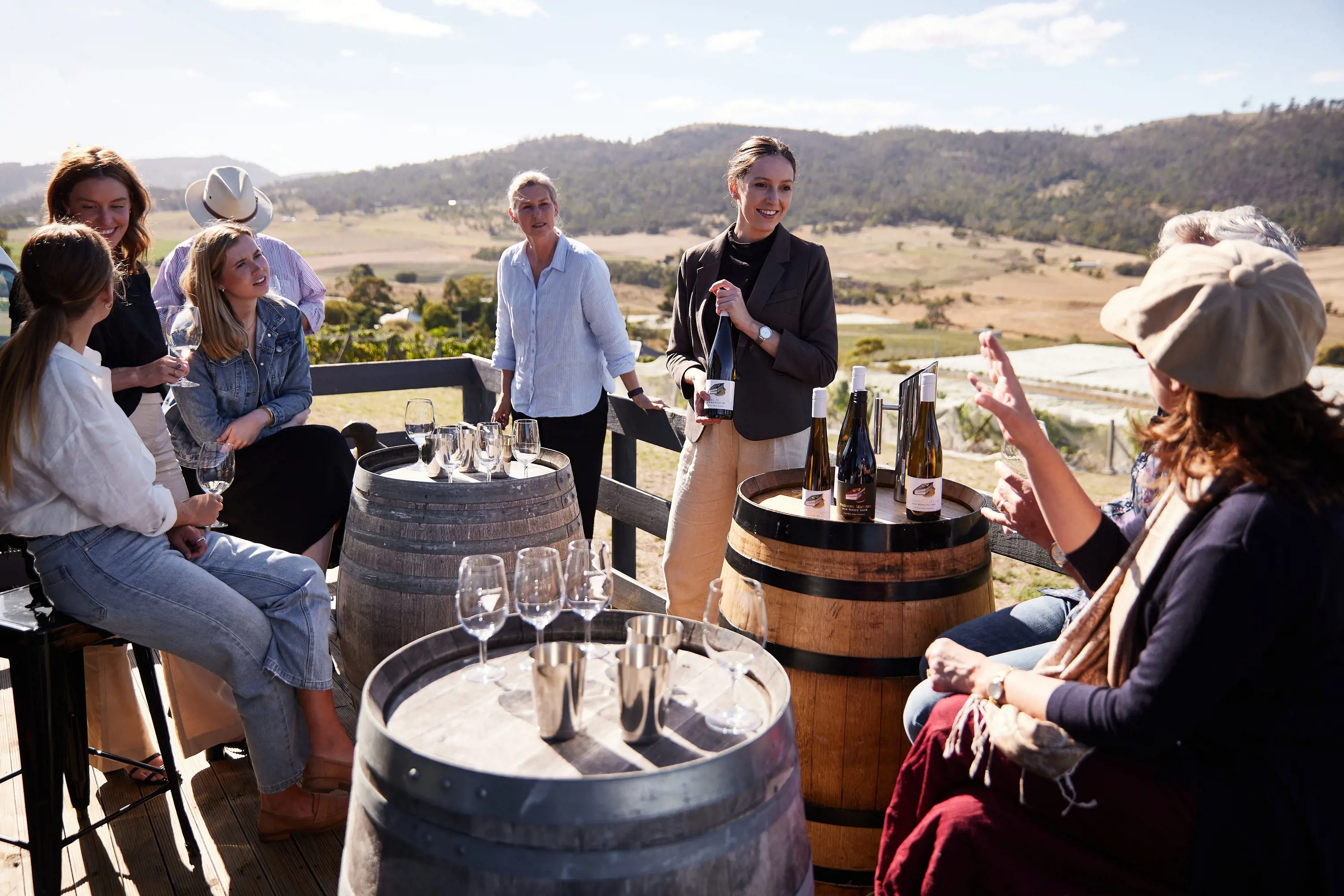 A group of people sit around up-turned barrels as a sommelier describes the wine they are drinking.