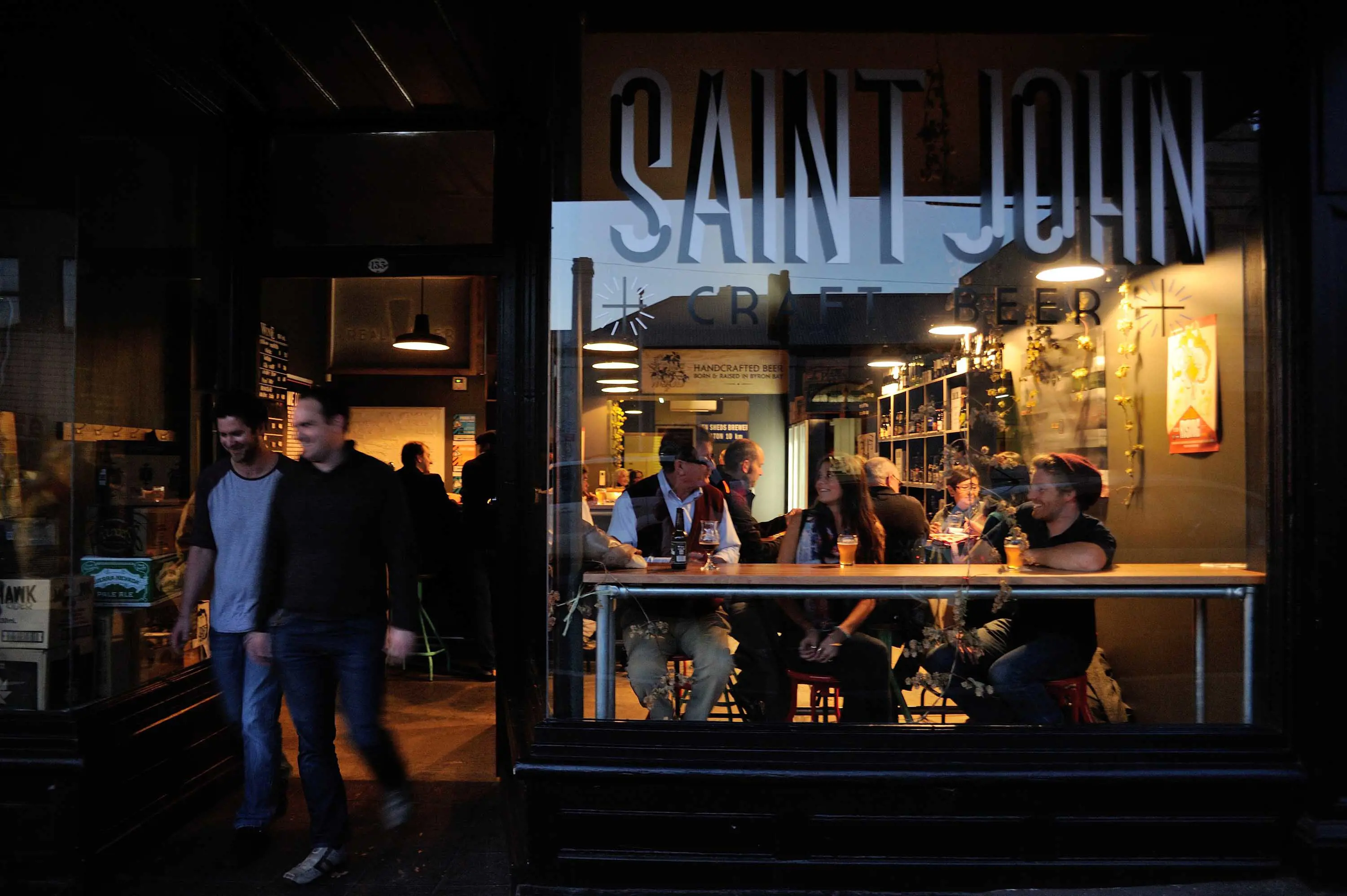 Two young men leave the entrance of a craft beer bar. An elderly man is sitting in the window at a high bench, talking with other customers.