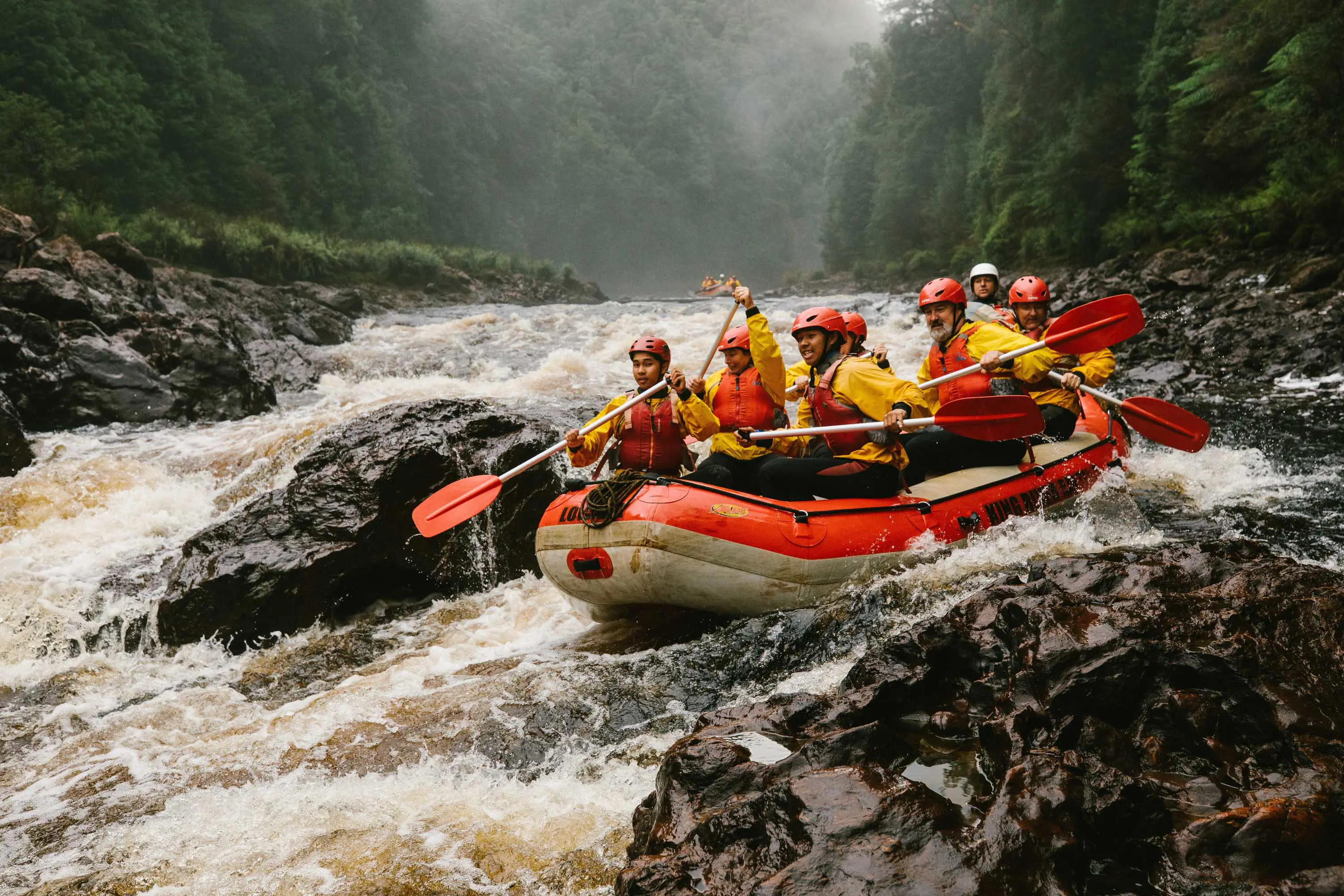 A group of rafters in brightly-coloured safety gear and helmets paddle their raft over fast moving rapids between rocks. 