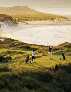 A couple tee off at a hole overlooking the white sands of a beach.