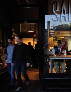 Two young men leave the entrance of a craft beer bar. An elderly man is sitting in the window at a high bench, talking with other customers.