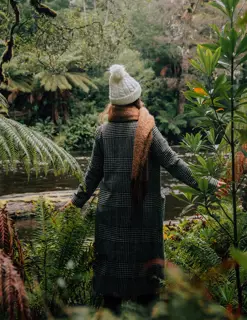 A woman wearing a white, woolen beanie and a winter coat stands in ferns and looks across a river into the forest.