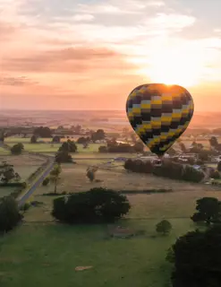 A large air balloon floats above green fields at sunrise.