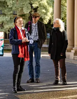 A tour guide speaks to a small group of people from the street in front of a large sandstone building with roman style pillars.