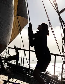 A woman stands in silhouette, pulling at a rope securing sails on a large sailing boat.