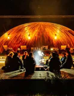 A group of people sit around a fire pit in front of a modern, illuminated hut during darkness. 